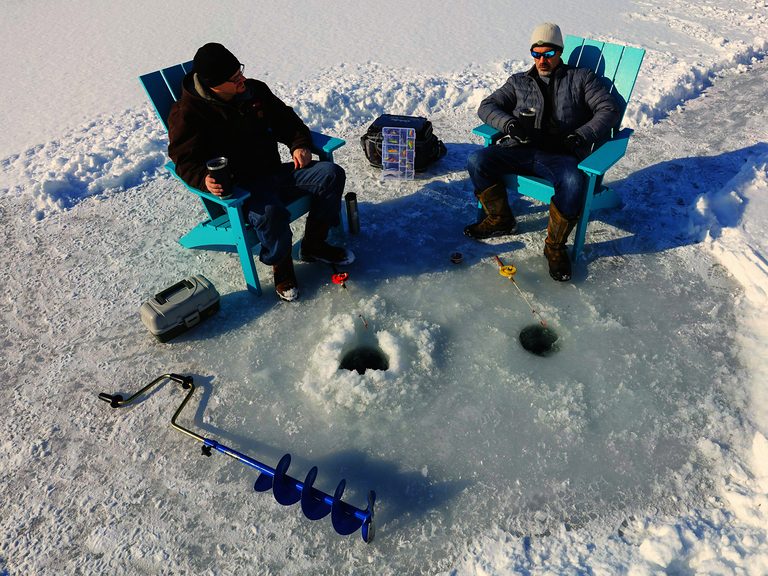 Two anglers sitting in chairs on a frozen lake beside drilled ice holes and a hand auger