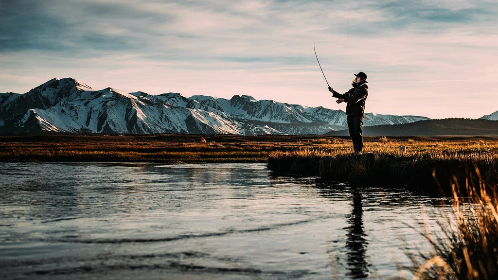 An angler casting a fly line on a still mountain river at golden hour, snow-dusted peaks behind