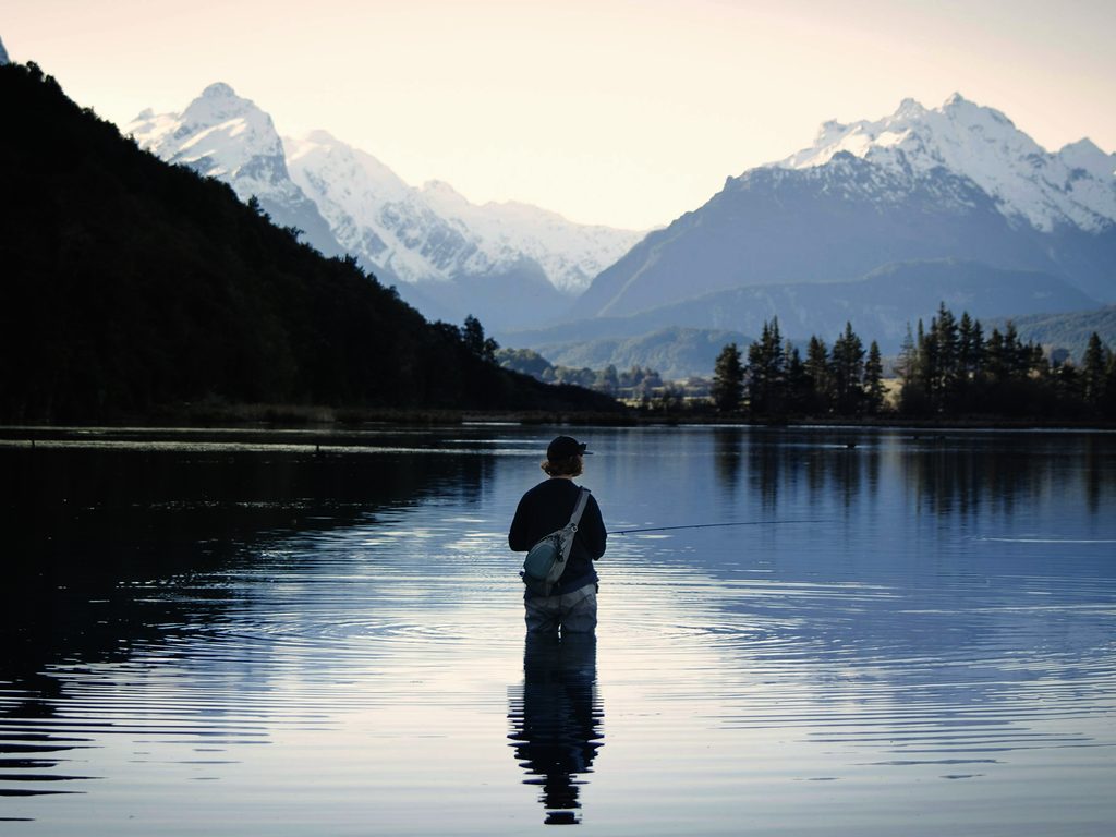 A lone angler standing waist-deep in glassy still water beneath misty coastal mountains